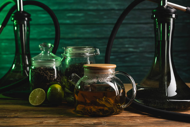 Teapot near blurred limes and hookahs on wooden table on wooden background  - Valokuva, kuva
