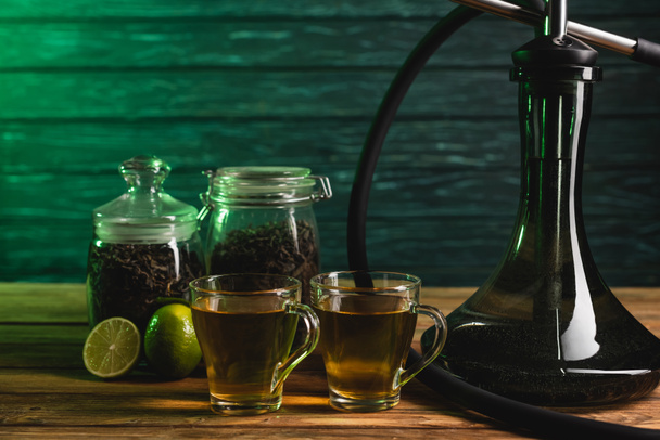 Cups of tea, limes and hookah on wooden surface on wooden background  - Photo, Image