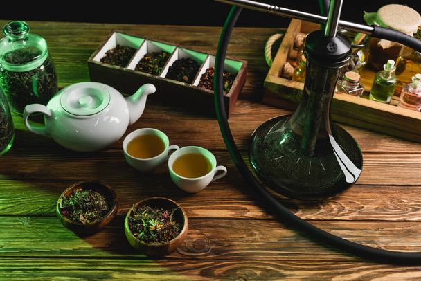 Top view of teapot, dry tea and hookah on wooden surface isolated on black  - Photo, Image