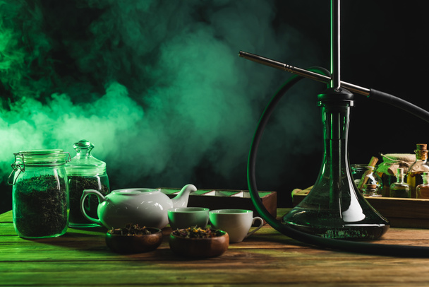 Hookah near dry tea and teapot on wooden surface on black background with smoke - Fotó, kép