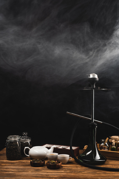 Cups of tea, hookah on wooden surface on black background with smoke - Photo, Image