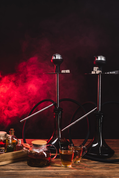 Cups of tea near teapot and hookahs on wooden surface on black background with red smoke  - Фото, зображення