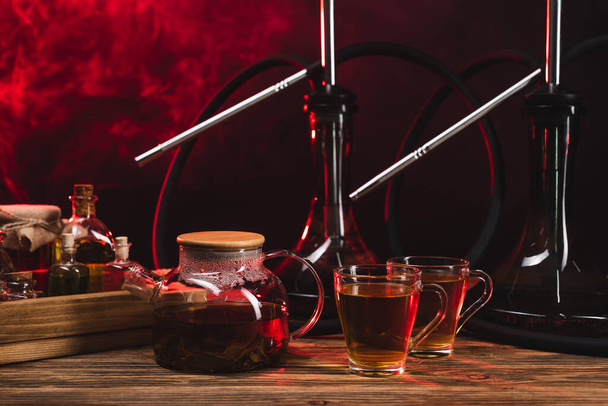 Tee in der Nähe von Wasserpfeifen auf hölzerner Oberfläche auf schwarzem Hintergrund mit Rauch  - Foto, Bild