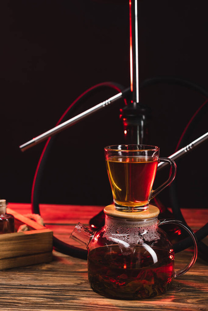 Cup of tea on teapot near hookah on wooden surface isolated on black - Photo, Image