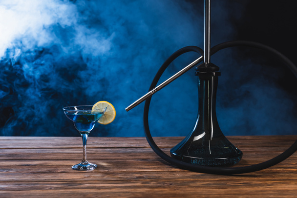 Glass with cocktail near hookah on wooden surface on black background with smoke - Photo, Image