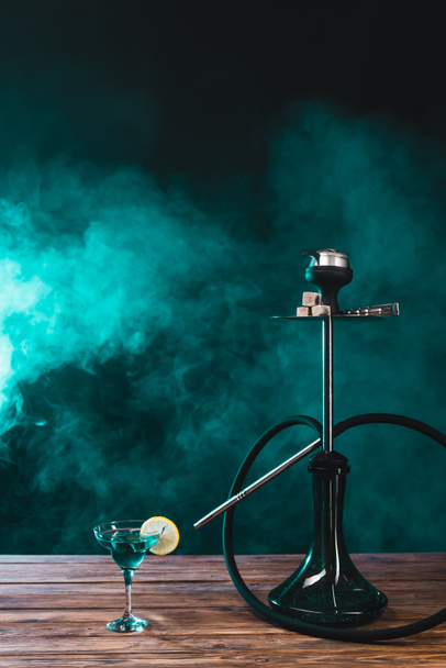 Hookah near glass of cocktail on wooden surface on black background with green smoke - Foto, imagen