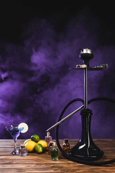 Narghilè vicino cocktail su superficie di legno su sfondo nero con fumo viola - Foto, immagini