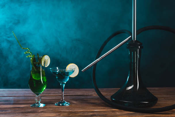 Glass of cocktails near hookah on wooden surface on black background with green smoke - Photo, Image