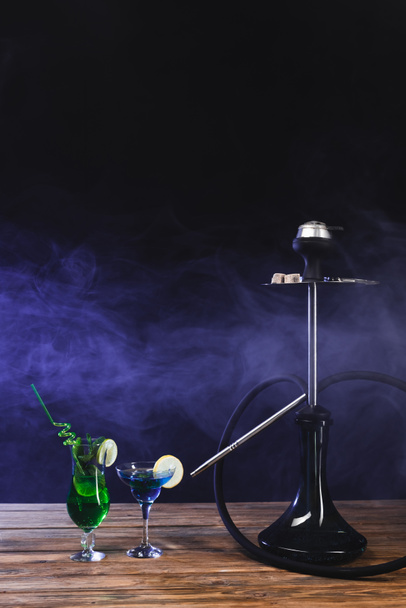 Hookah near glasses of cocktails on wooden surface on black background with smoke - 写真・画像