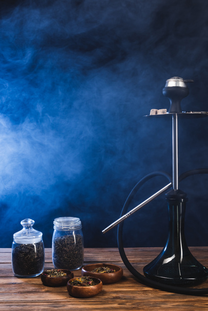 Hookah near dry tea in bowls on wooden surface on black background with blue smoke - Photo, Image