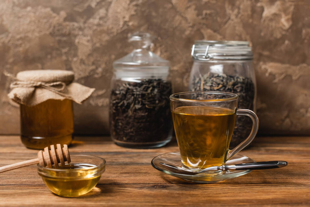 Cup of tea near honey and blurred jars on wooden surface on textured stone background  - Foto, Bild