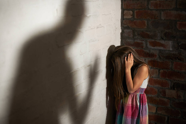 Wehrloses Opfer. Misshandeltes kleines Mädchen zusammengekauert, während der Schatten ihres Täters auf sie zukommt. - Foto, Bild