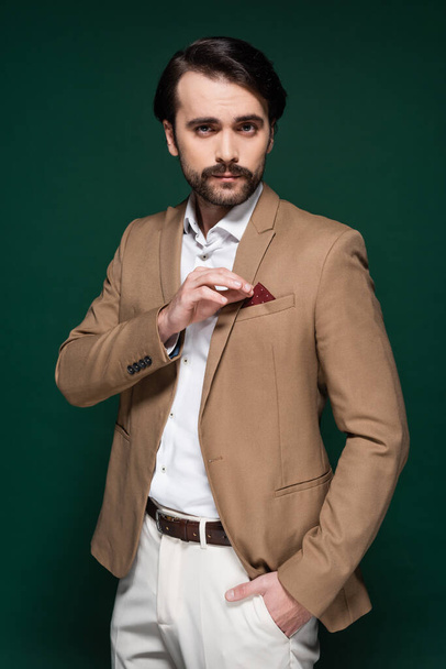 young man with mustache adjusting handkerchief in pocket of beige blazer on dark green - 写真・画像