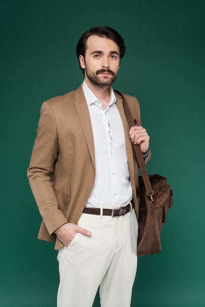 man with mustache standing with hand in pocket and holding leather bag on dark green  - Φωτογραφία, εικόνα