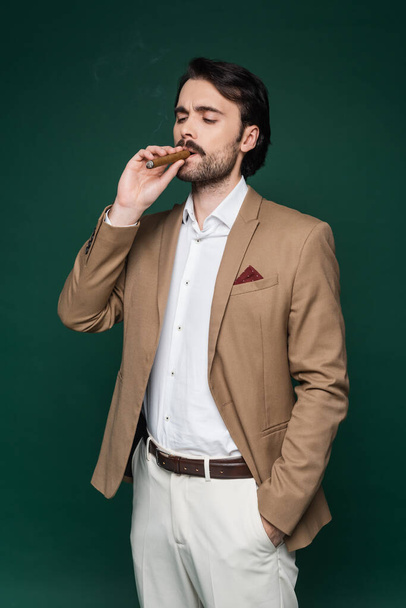 man with mustache smoking cigar and standing with hand in pocket on dark green  - Foto, Imagen