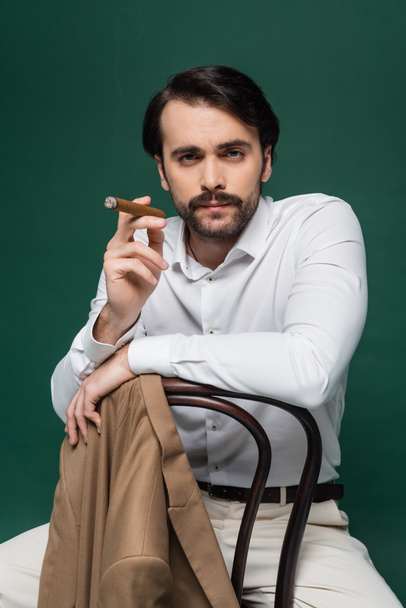 man with mustache holding cigar and sitting on chair on dark green  - Фото, зображення