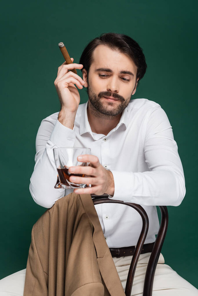 man with mustache holding cigar and glass of brandy while sitting on chair on dark green  - Photo, Image