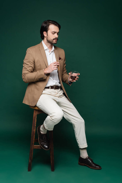 full length of man in blazer holding cigar and glass of whiskey while sitting on chair on dark green  - Foto, imagen