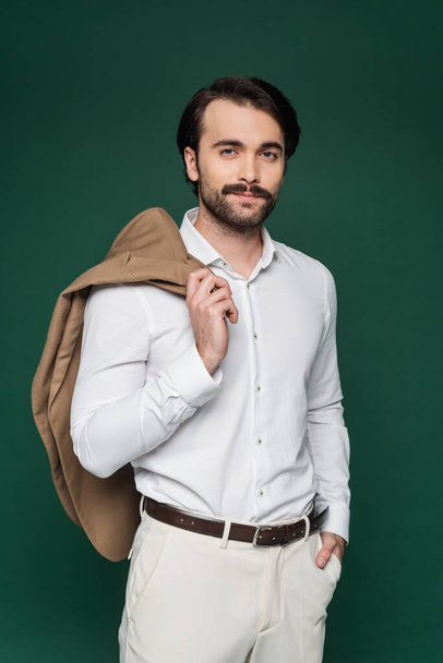 man with mustache holding beige blazer and standing with hand in pocket on dark green  - Foto, immagini