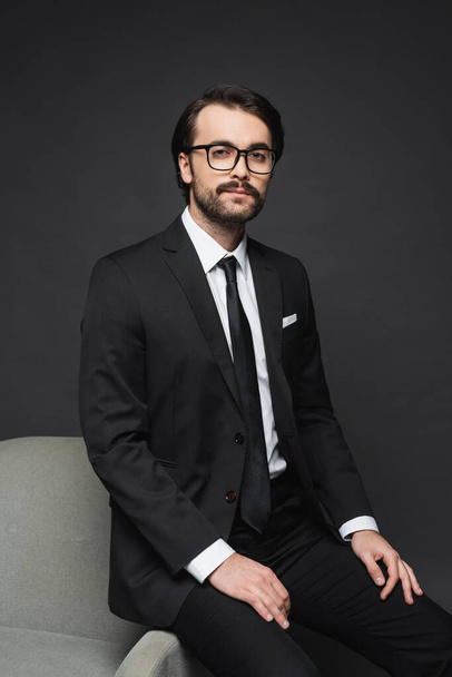 businessman in formal wear and glasses leaning on armchair on dark grey - Фото, изображение