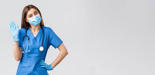 Covid-19 、ウイルス、健康、医療従事者および検疫の概念を防止する。青いスクラブと医療マスクを身に着けているクリニックでフレンドリーな女性看護師,手袋でこんにちはやハイサインを振ってオン - 写真・画像