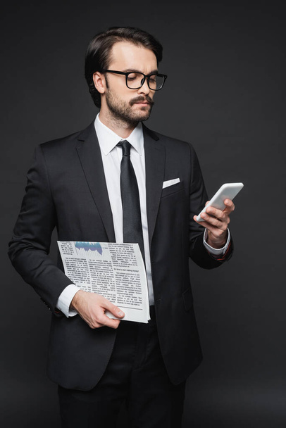 businessman in suit and glasses holding newspaper and using smartphone on dark grey - Foto, afbeelding
