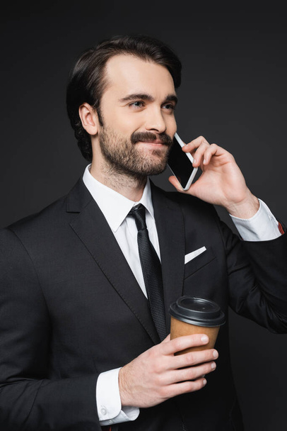 smiling businessman with mustache holding paper cup and talking on smartphone on dark grey - Φωτογραφία, εικόνα