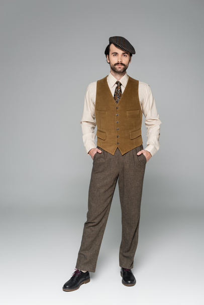 volledige lengte van de man met snor en retro kleding poseren met de handen in zakken op grijs - Foto, afbeelding