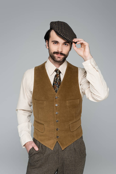 man met snor en retro kleding poseren met de hand in zak terwijl het aanpassen van vintage hoed geïsoleerd op grijs - Foto, afbeelding