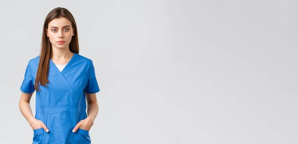 Healthcare workers, prevent virus, insurance and medicine concept. Serious-looking, confident young medical worker, nurse or doctor in blue scrubs hold hands in pockets and looking camera - Fotoğraf, Görsel