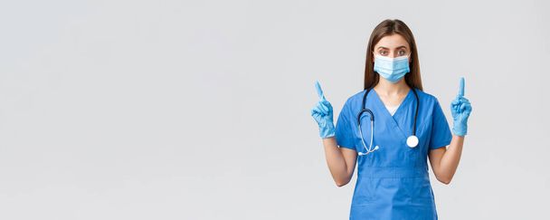 Ковід-19, запобігання вірусу, здоров'ю, концепції працівників охорони здоров'я. Серйозна і впевнена жіноча медсестра в блакитних скрабах, медична маска PPE, спрямована на пальці вгору, інформує пацієнтів, як запобігти зараженню корони
 - Фото, зображення