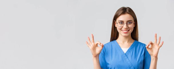 Медичні працівники, медицина, страхування та концепція пандемії ковадла-19 . Впевнена та оптимістична медсестра-жінка, лікарка в блакитних скрабах та окулярах, демонструє правильний знак, гарантує найкращу якість обслуговування
 - Фото, зображення