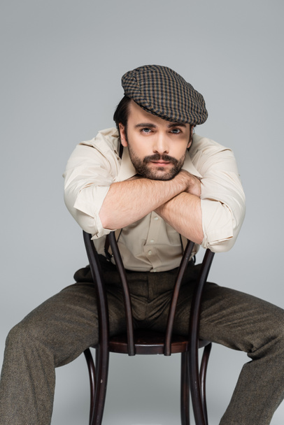 man with mustache in retro style clothing and hat sitting on wooden chair on grey  - Foto, Imagem