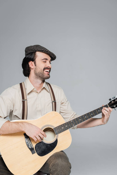 homme heureux avec moustache dans des vêtements de style rétro et chapeau jouant guitare acoustique isolé sur gris  - Photo, image