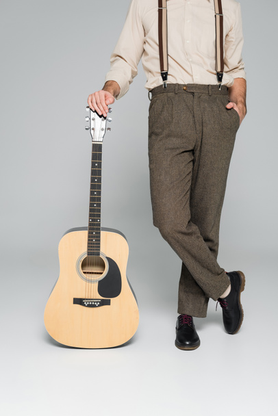 vista cortada do homem de pé com a mão no bolso perto da guitarra acústica em cinza  - Foto, Imagem