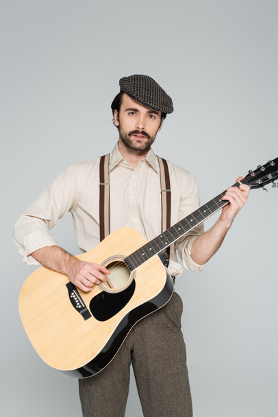 άνδρας με μουστάκι σε ρετρό ρούχα και καπέλο παίζοντας ακουστική κιθάρα απομονωμένος σε γκρι  - Φωτογραφία, εικόνα