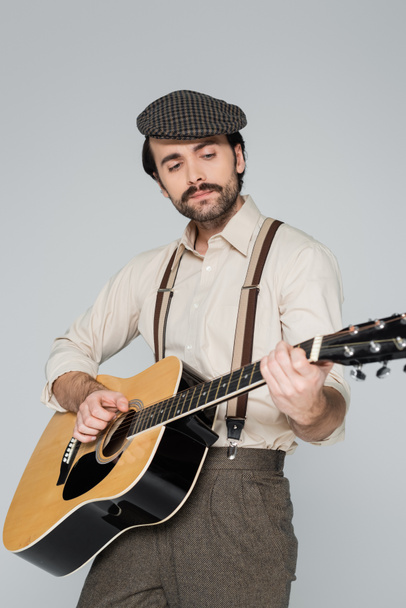 jeune homme avec moustache dans des vêtements de style rétro jouant de la guitare acoustique isolée sur gris  - Photo, image