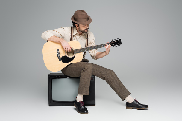 full length of man retro style clothing playing acoustic guitar and sitting on antique tv on grey - Photo, Image