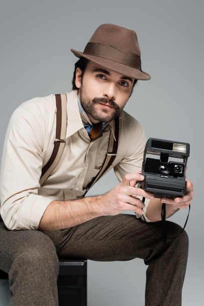 man retro style clothing and hat holding vintage camera and sitting on antique tv isolated on grey  - Photo, Image