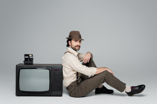 full length of man retro style clothing and hat sitting near antique tv with vintage camera on grey  - Φωτογραφία, εικόνα
