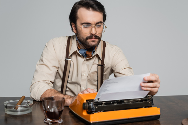 periodista en anteojos mirando papel en máquina de escribir cerca de vaso de whisky y cigarro aislado en gris - Foto, Imagen