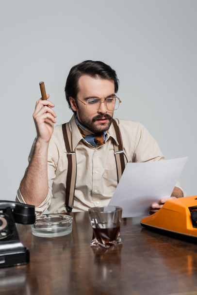 periodista con bigote mirando el papel y sosteniendo cigarros cerca de la máquina de escribir y un vaso de whisky en el escritorio aislado en gris - Foto, Imagen