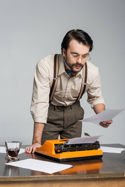 journalist in eyeglasses looking at paper near typewriter on desk isolated on grey - Фото, изображение