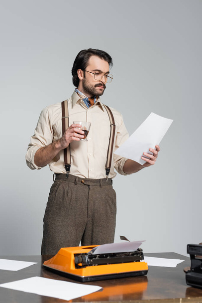 dziennikarz w okularach patrzący na papier i trzymający szklankę whisky w pobliżu maszyny do pisania na biurku odizolowany na szaro - Zdjęcie, obraz