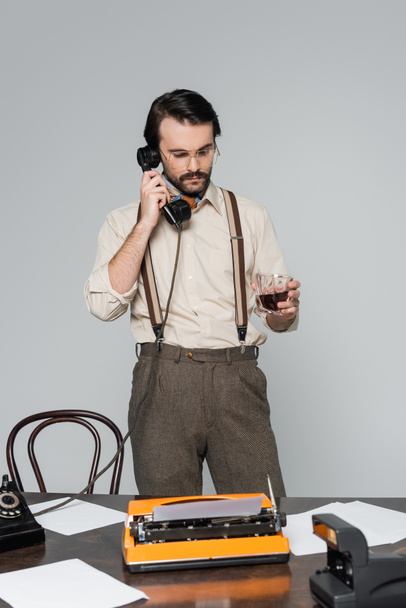 periodista en gafas con vaso de whisky cerca de la máquina de escribir en el escritorio y hablando por teléfono retro aislado en gris - Foto, Imagen