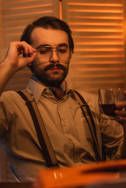 Gözlükleri ayarlayan ve bulanık perdenin yanında viski bardağı tutan adam. - Fotoğraf, Görsel