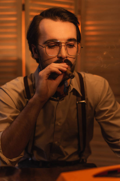 man with mustache in eyeglasses smoking cigar near blurred folding screen - 写真・画像