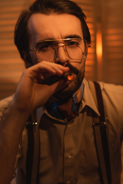 άνδρας με γυαλιά καπνίζοντας πούρο και κοιτάζοντας την κάμερα κοντά σε θολή πτυσσόμενη οθόνη - Φωτογραφία, εικόνα