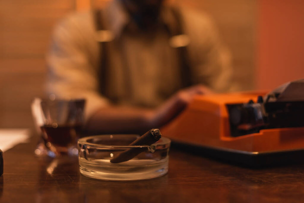 Aschenbecher mit Zigarre neben Schreibmaschine und verschwommenem Mann im Hintergrund  - Foto, Bild