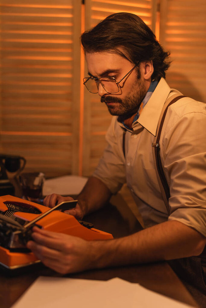 man with mustache and eyeglasses looking at retro typewriter machine on desk - Foto, Imagem